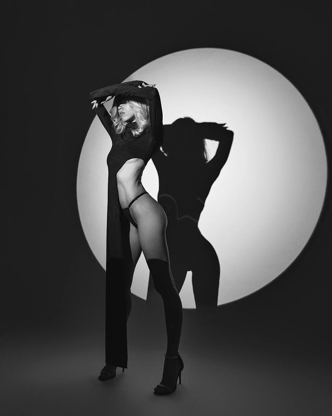 Woman in black lingerie and high heels posing with her arms above her head, creating a shadow on the wall behind her against a circular backdrop.