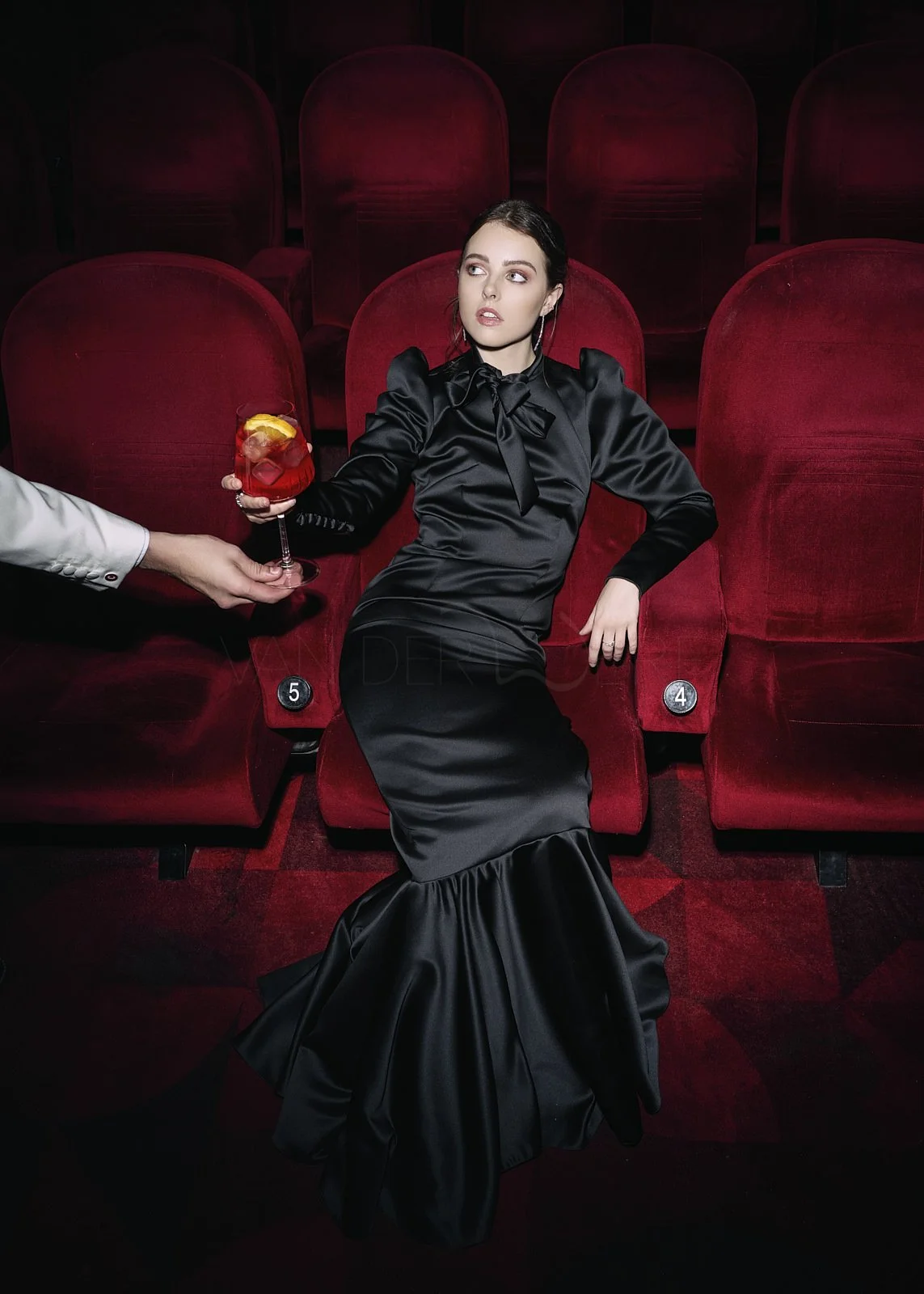 A woman in a black satin gown sitting in red theater seats, holding a glass of red drink with lemon. A person in a white jacket is serving her the drink. The woman looks to the side with a neutral expression.