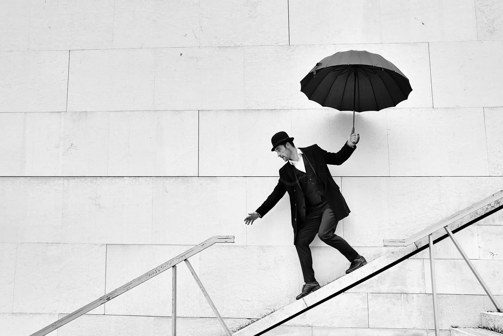 A man in a suit and bowler hat holding an umbrella while walking down a staircase in front of a plain wall.