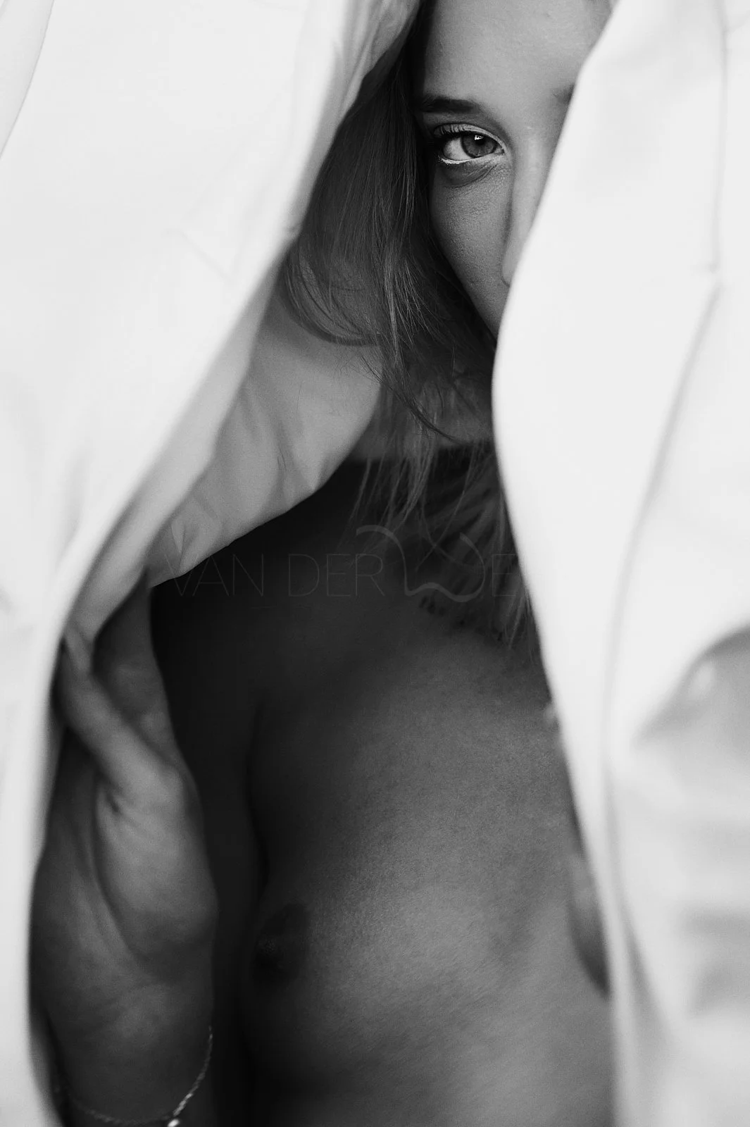 A black and white photo of a partially undressed woman with long hair, peering out from behind curtains or sheets, with one eye visible.