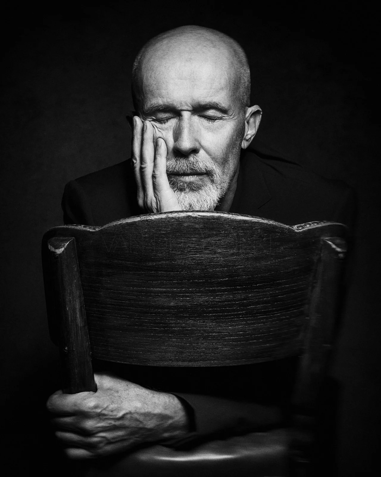 Black and white photo of a man with closed eyes, resting his face on his hand, sitting behind a wooden chair with his arm over the top.