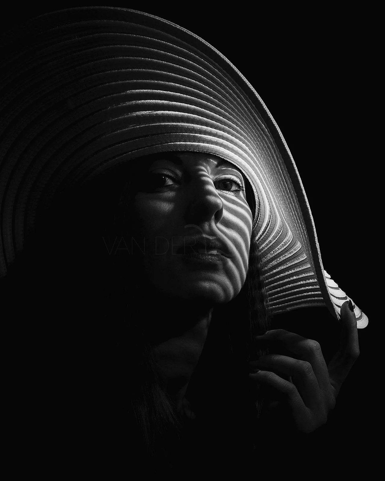 Black and white portrait of a woman wearing a wide-brimmed hat, with light highlighting her facial features and the hat's texture, against a dark background.