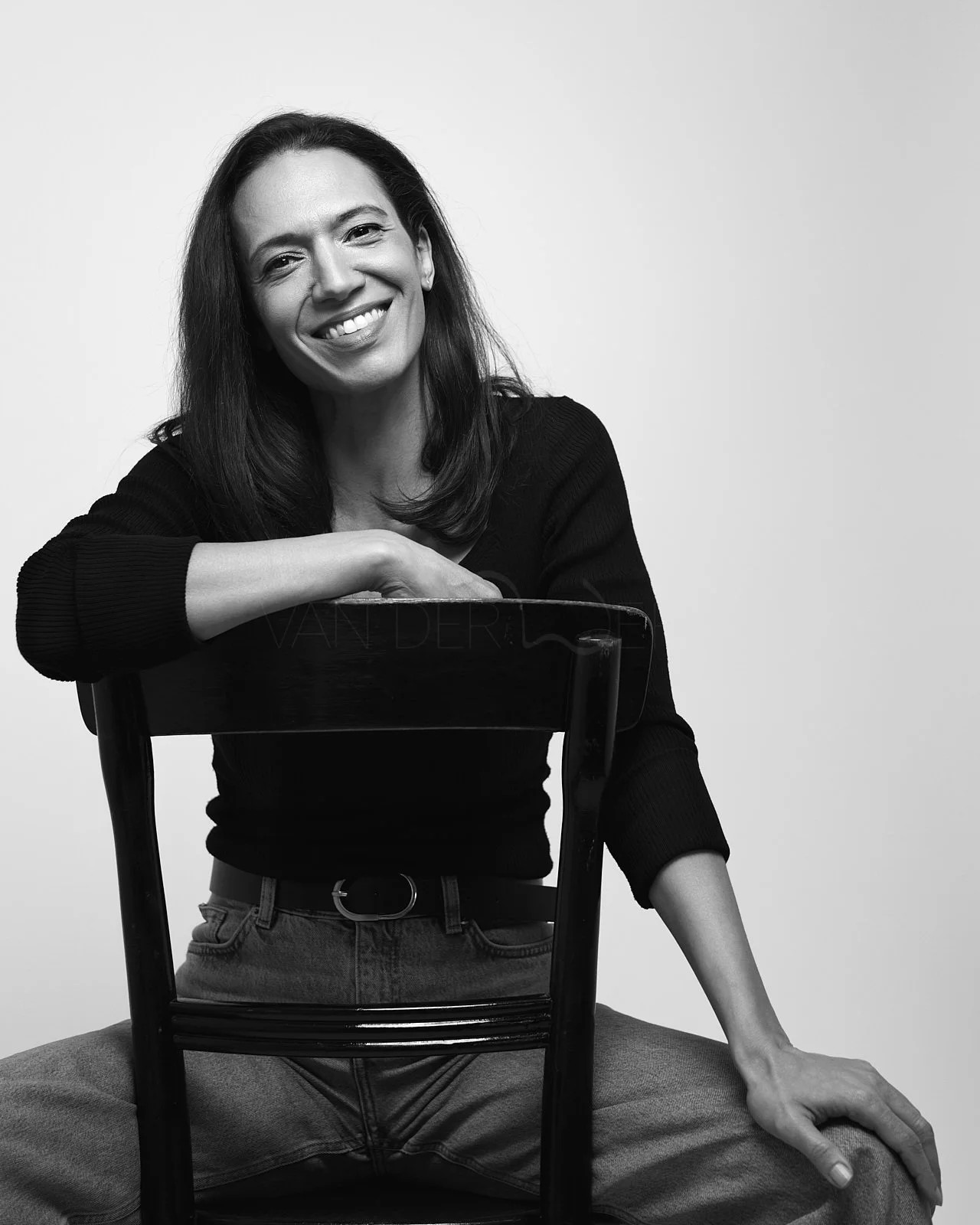 Black-and-white photo of a woman with dark hair, wearing a black long-sleeve top and jeans, smiling and sitting on the floor with one knee up, leaning on a chair with one arm resting on the chair back. The background is plain and light-colored.