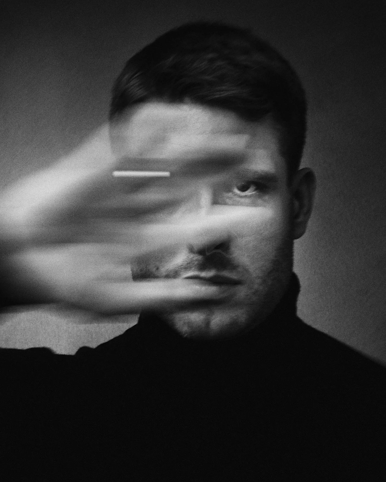 Black and white photo of a man with short hair, holding his hand in front of his face, partially obscuring it, with a blurred motion effect.