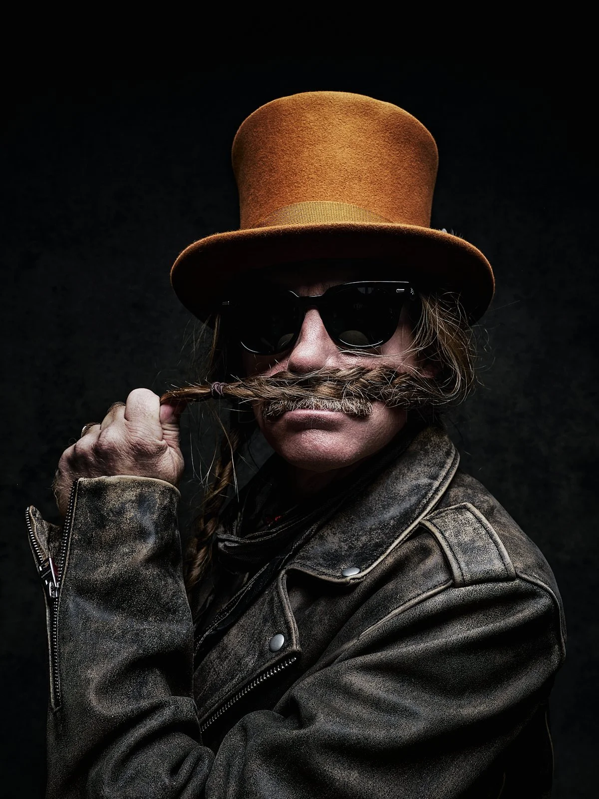 A person wearing a brown hat, sunglasses, and a leather jacket, holding a braided mustache to their face against a black background.