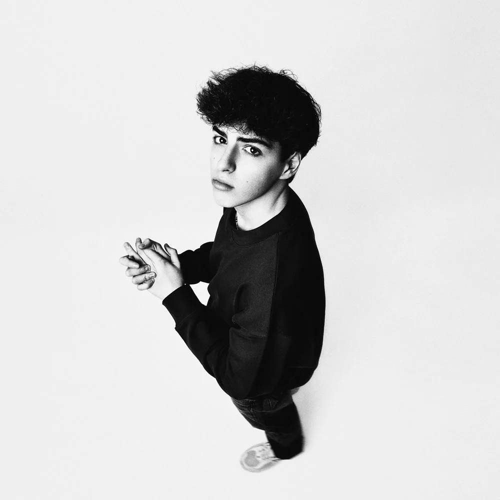 Black and white photo of a young man standing against a plain background, looking directly at the camera with hands clasped in front.