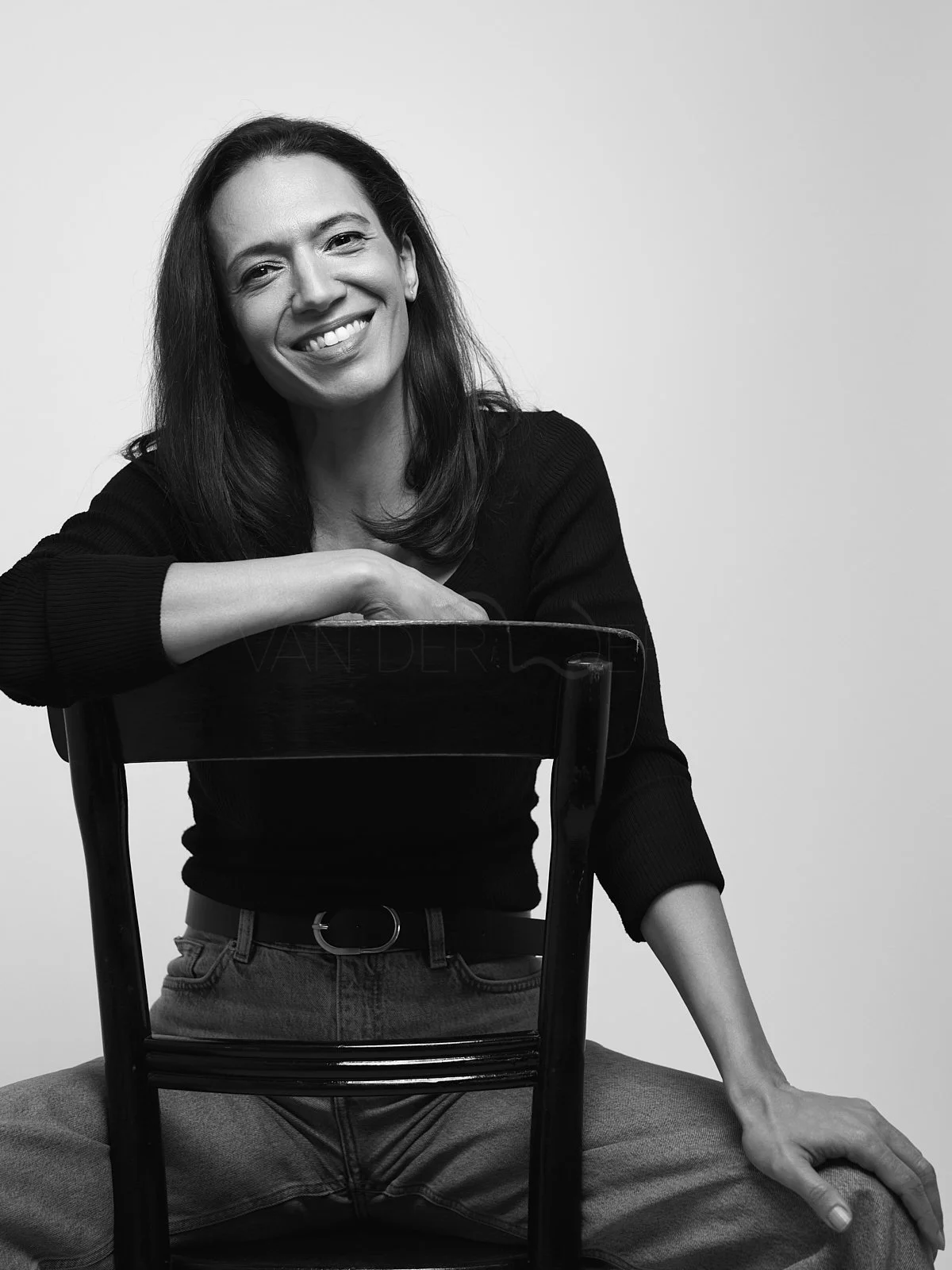 Black and white photo of a smiling woman with shoulder-length hair, sitting on the floor with her hands resting on a chair's backrest, wearing a dark long-sleeved sweater and jeans.