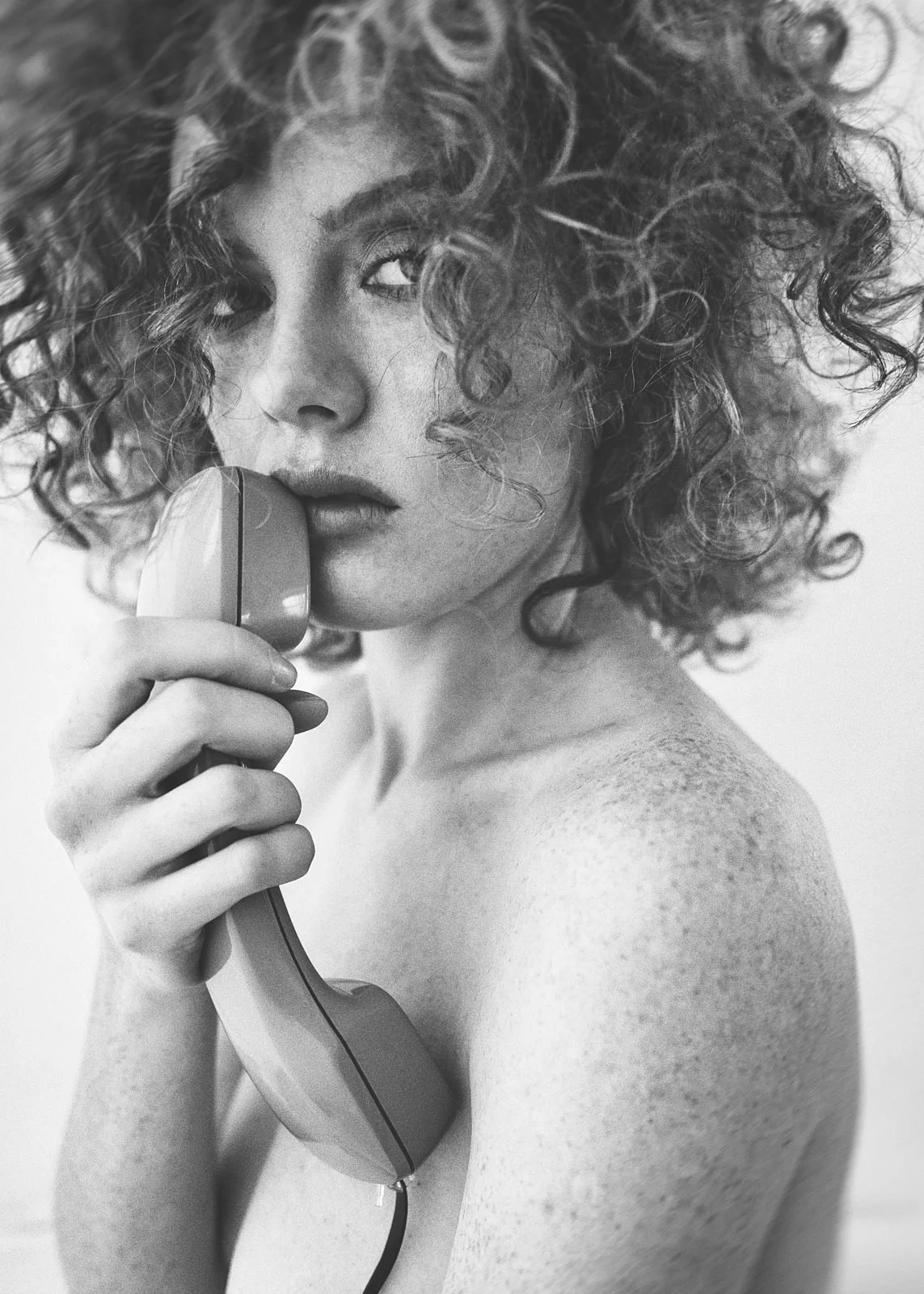 A black and white photo of a woman with curly hair holding a telephone receiver close to her lips, with her shoulder and part of her arm visible.