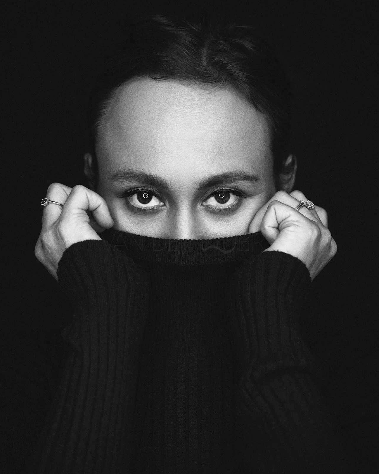 A black and white portrait of a woman with short dark hair, holding the collar of her black sweater up to her nose, revealing only her eyes and forehead against a dark background.