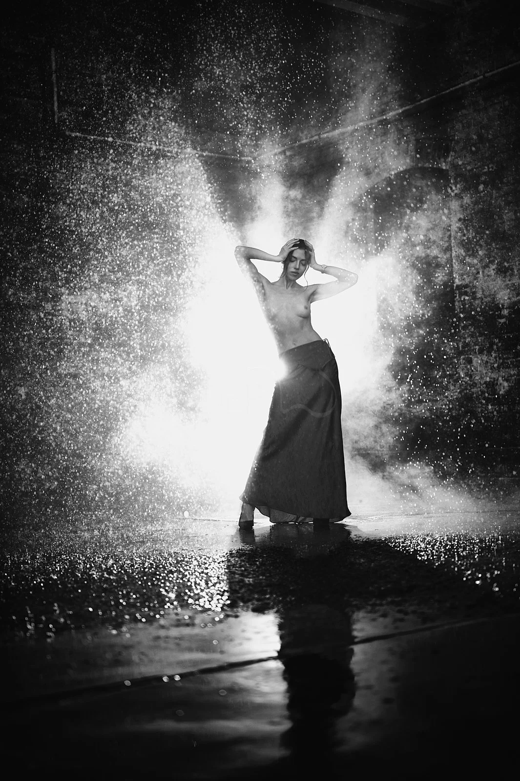 A woman standing in shallow water with water splashing and spraying around her, backlit creating a glowing effect, in a black and white photo.