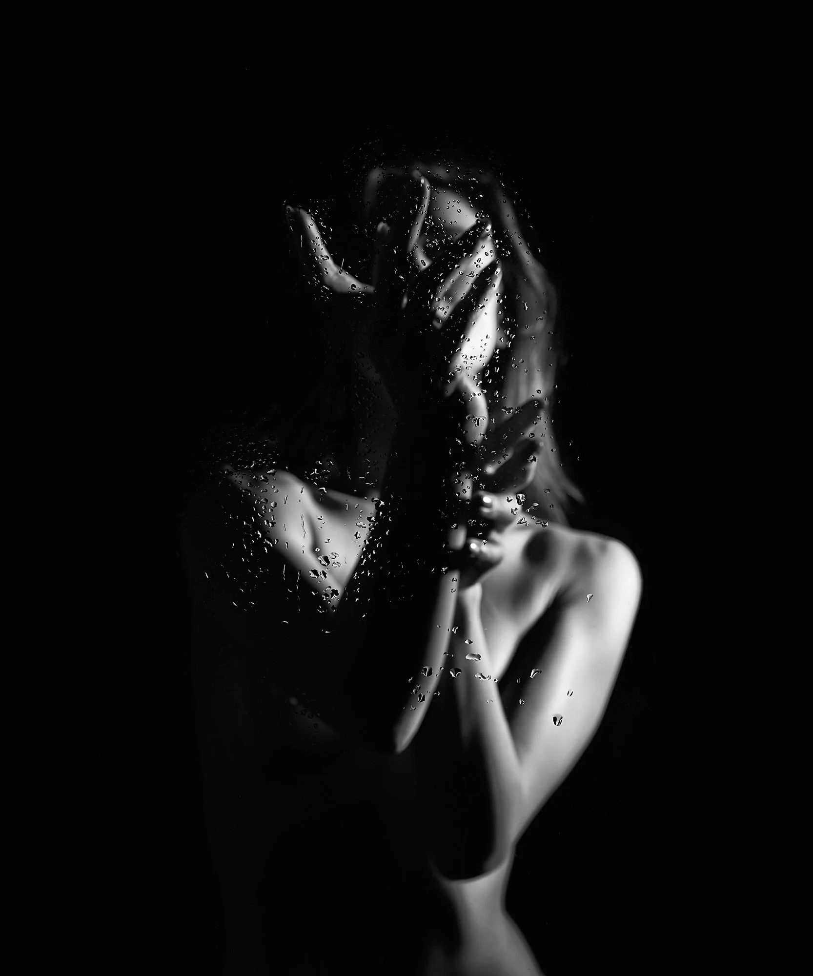 A black and white photo of a person with water droplets on a glass, covering their face and upper body, with hands held up to their face, obscuring their features.