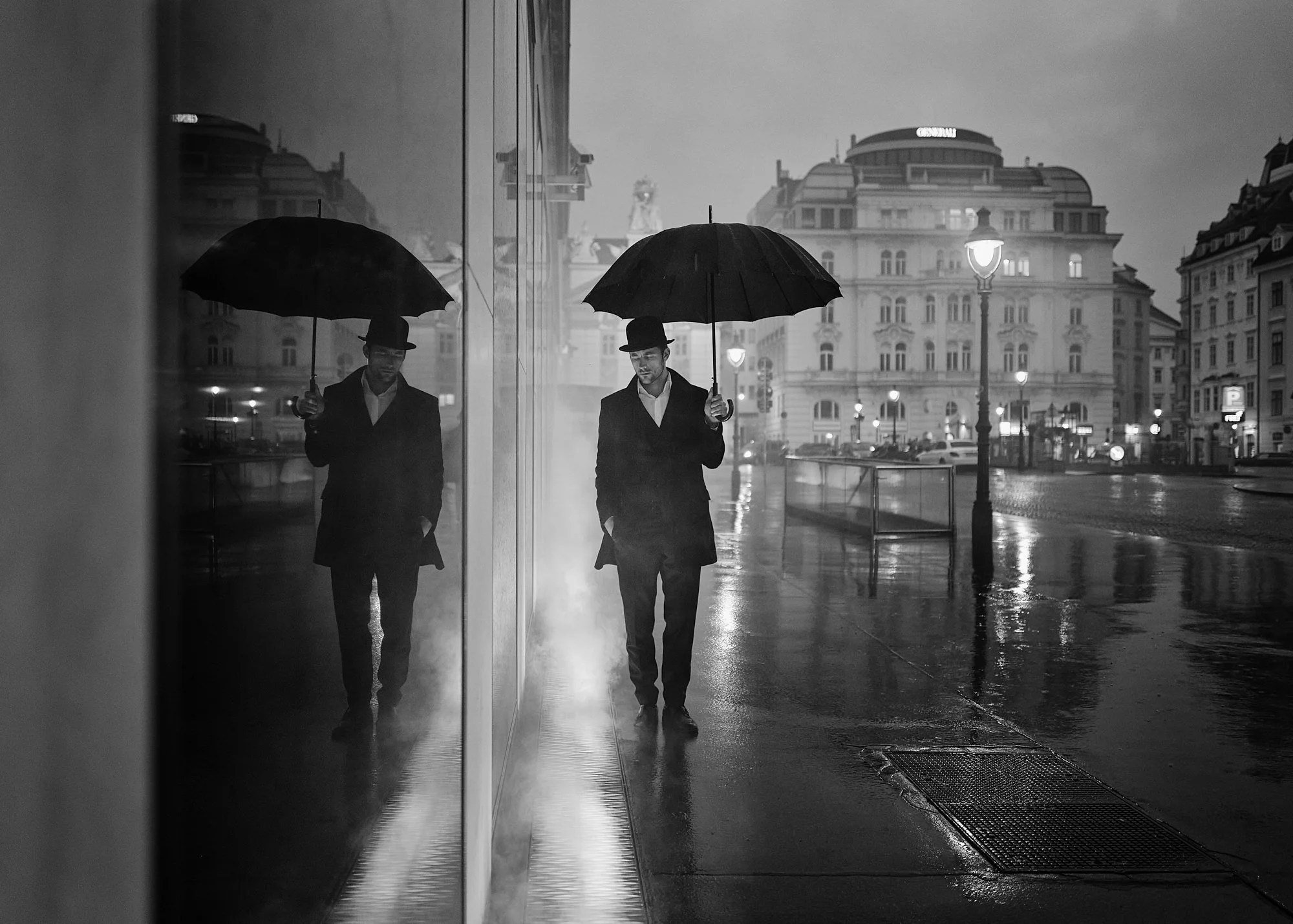 A man in a suit and hat holding an umbrella walking on a rainy city street at dusk, with his reflection visible in a glass building wall.