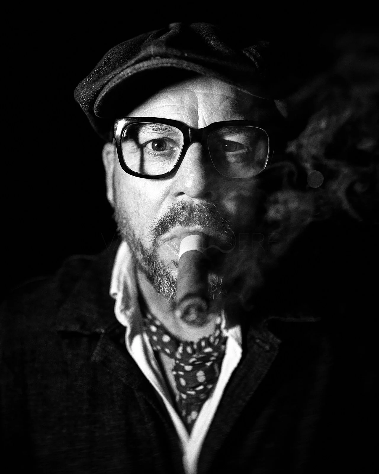 Black-and-white portrait of a man with glasses, a beard, wearing a hat, smoking a cigar, and wearing a polka-dot scarf.