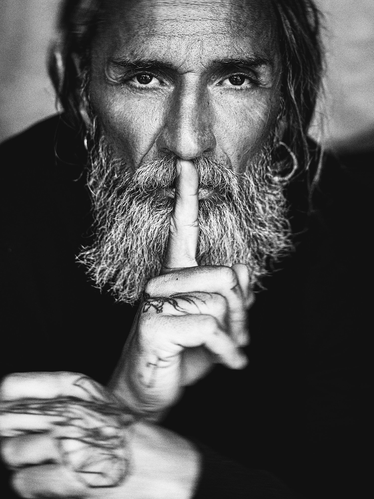 Black and white close-up of a bearded man with long hair holding his finger to his lips in a shushing gesture.