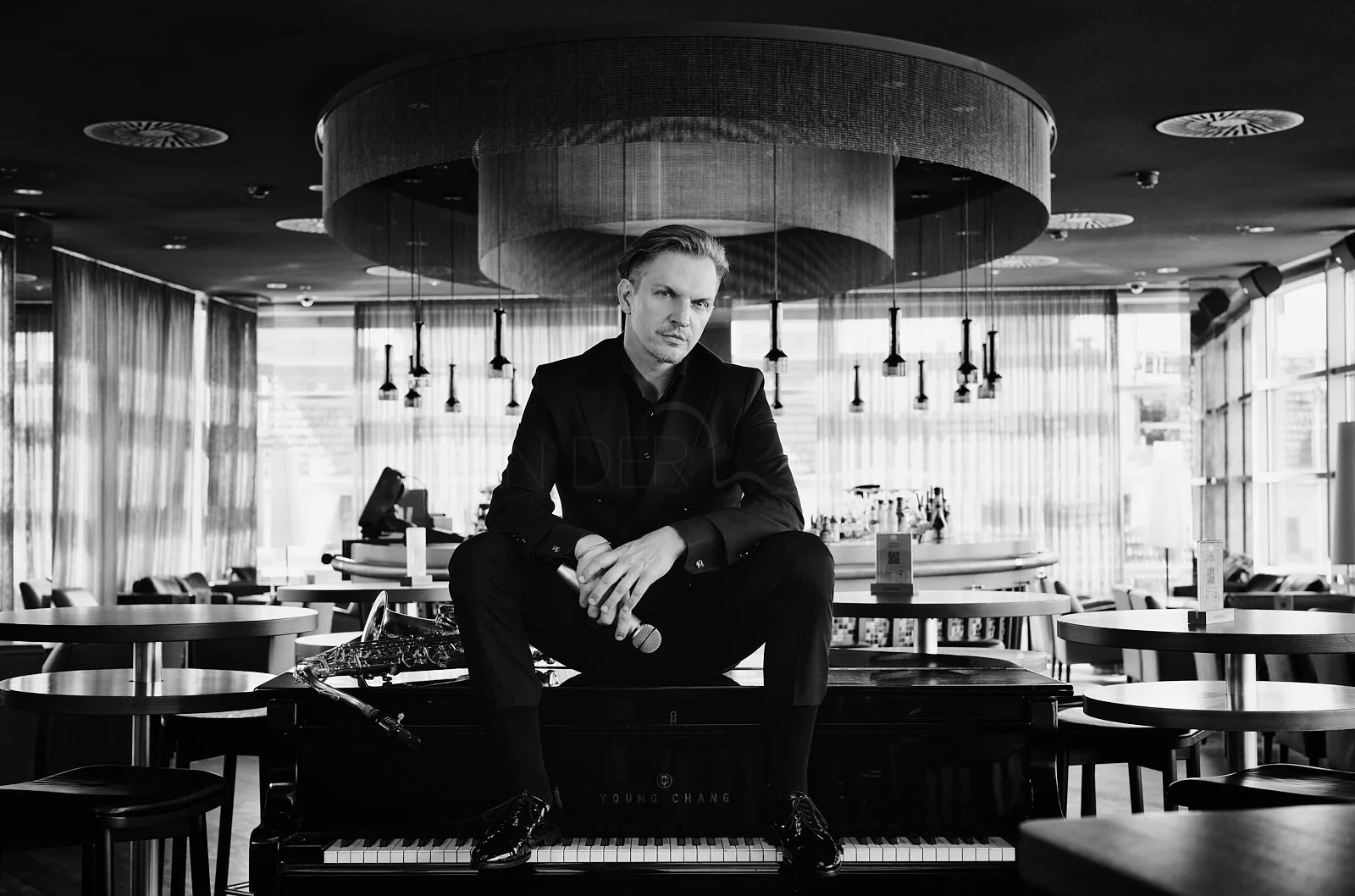 A man in a dark suit sitting on top of a piano in a modern, well-lit restaurant or café. He is holding a microphone in one hand and looking at the camera. The setting features round tables, chairs, large windows, and hanging pendant lights.