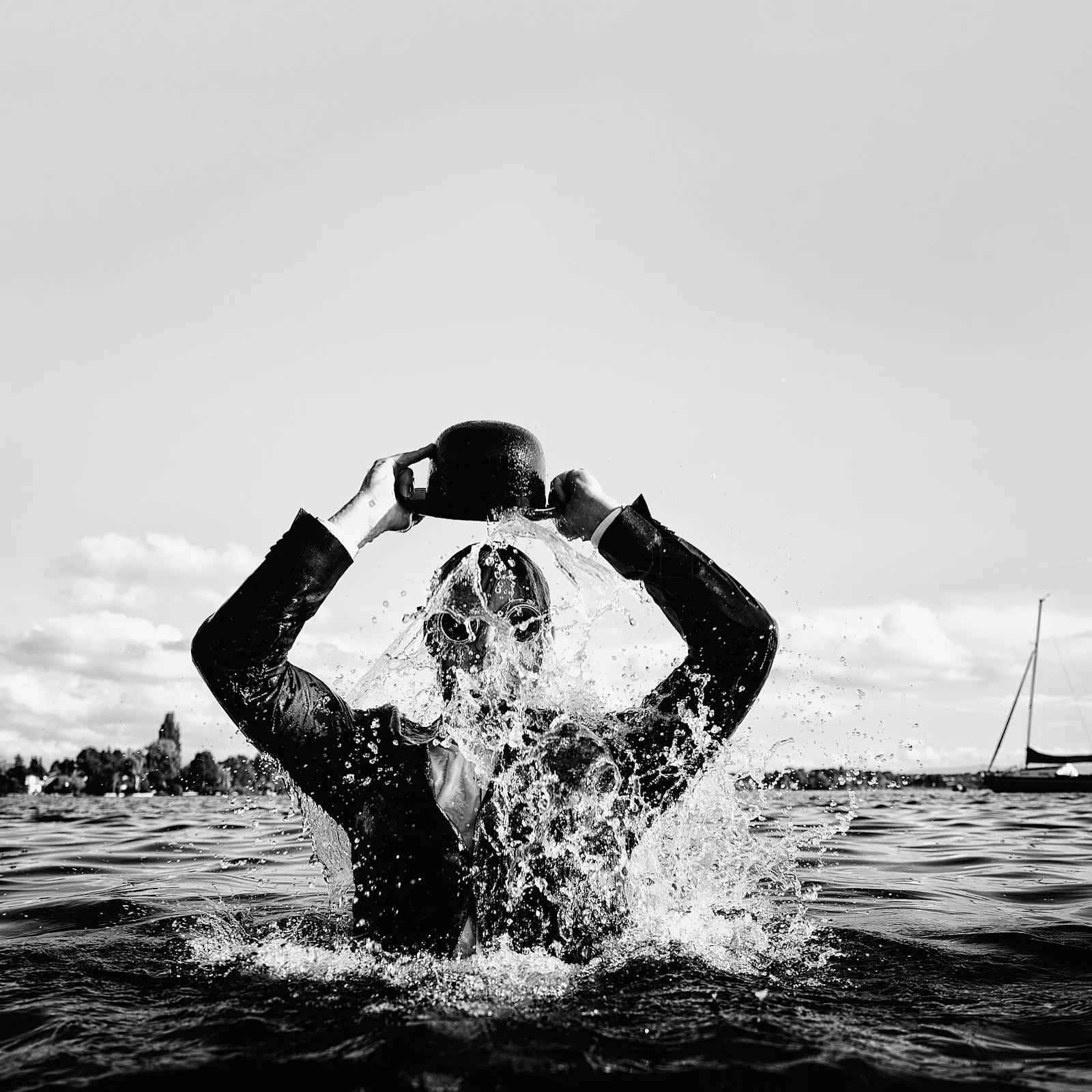 Person in a suit and swimming goggles holding a kettle above their head, pouring water over themselves in a body of water, black and white photo.