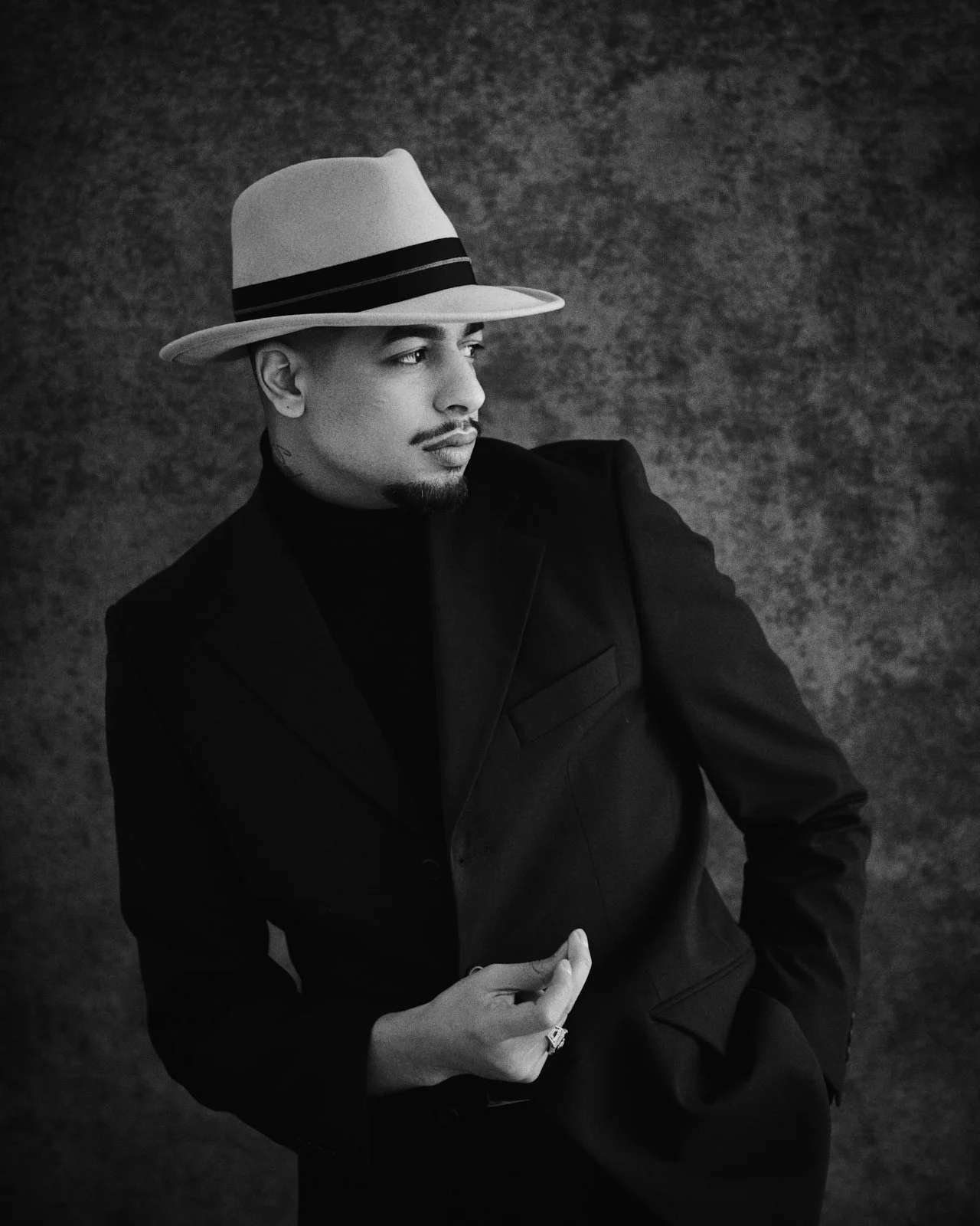 A black and white portrait of a man wearing a dark suit and a stylish fedora hat with a black band, posed against a textured background.