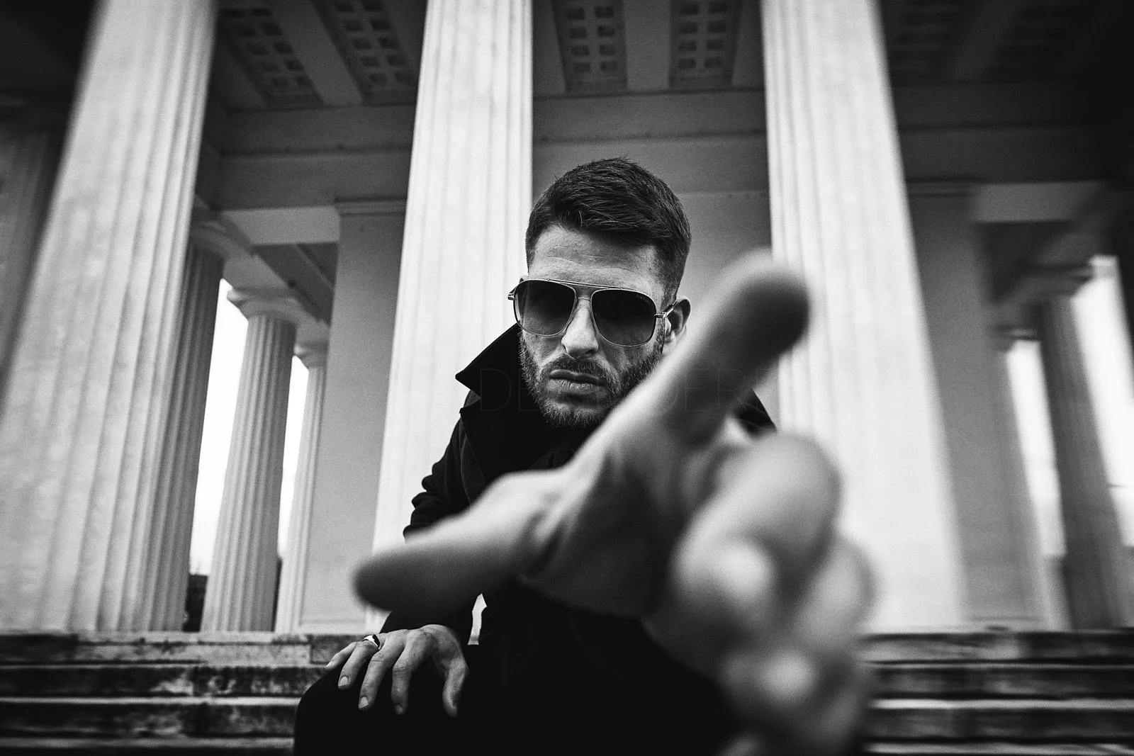 Black and white photo of a man wearing sunglasses and a dark jacket, leaning forward on the steps of a building with large columns, reaching toward the camera with his finger extended.