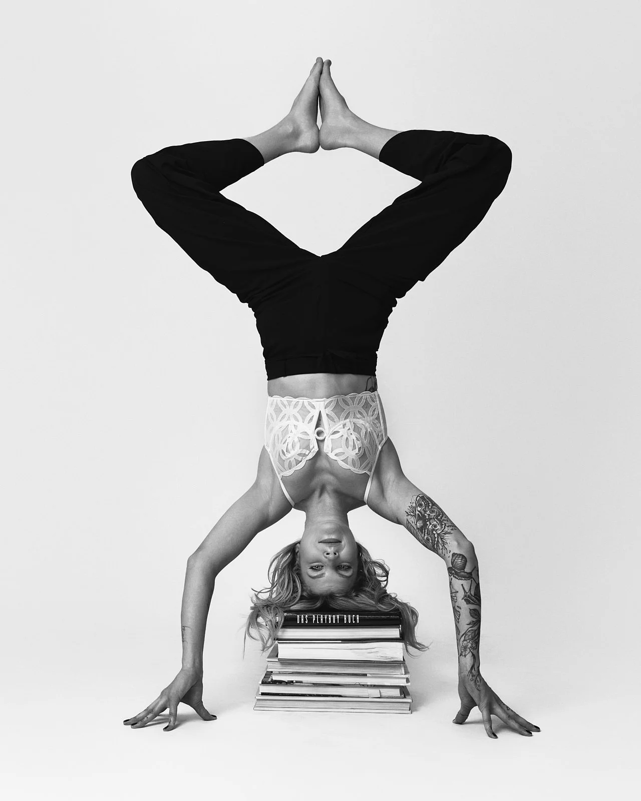 Woman performing a handstand with her head on a stack of books, her legs bent and hands touching at the palms above her head, in black and white.