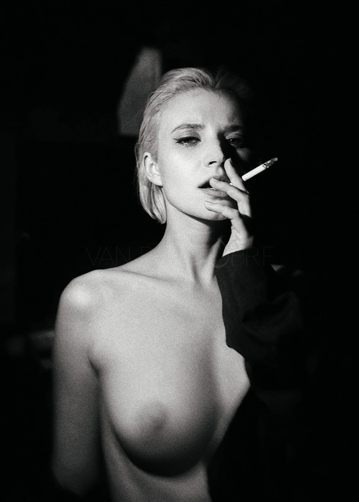 Black and white photo of a topless woman with short platinum hair holding a cigarette near her lips, with a high-contrast lighting and dark background.