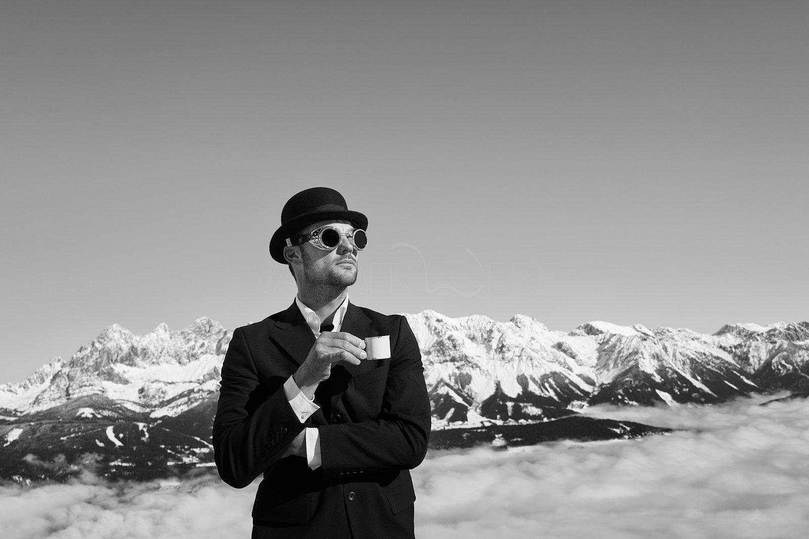 Man in vintage attire with goggles and bowler hat, holding a tea cup, standing in front of snow-capped mountains and clouds.