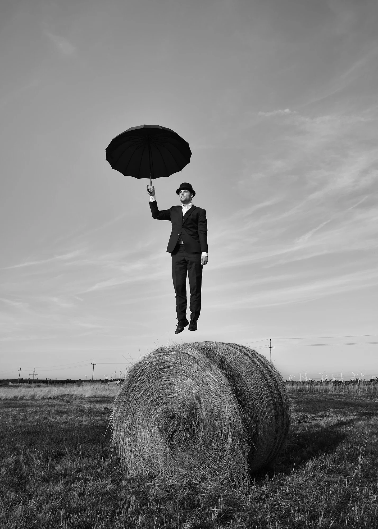 A man in a suit and bowler hat appears to be floating above a hay bale in a field, holding an umbrella. The image is in black and white.