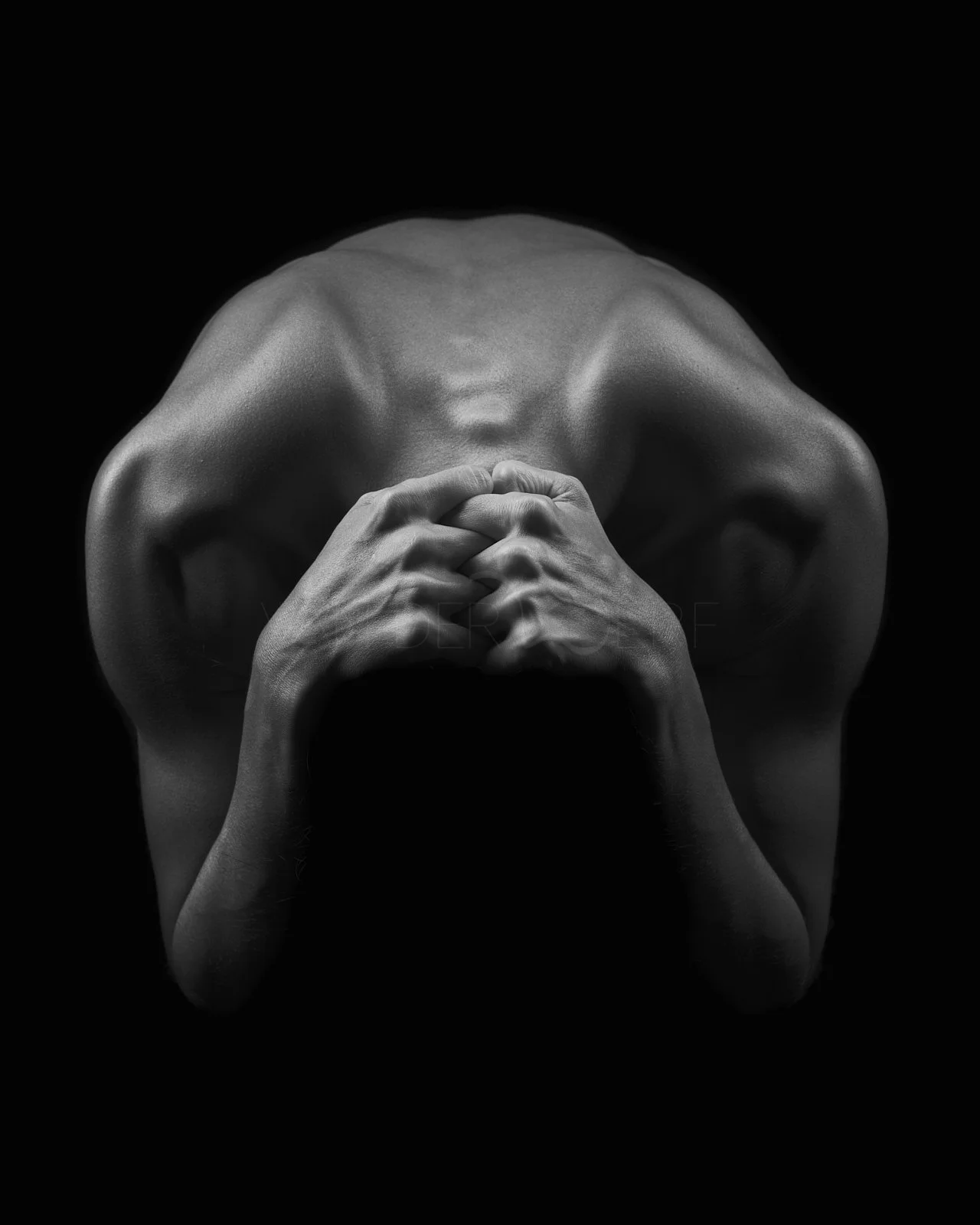 Black and white photo of a nude person with flexed muscles, head bowed, hands clasped, against a black background.