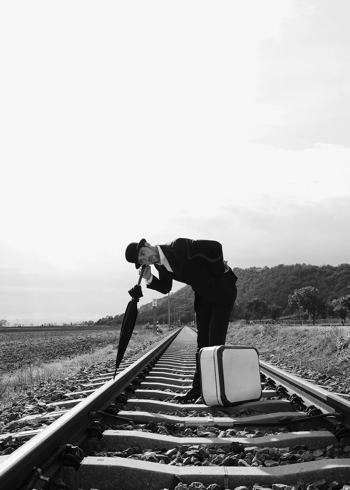 A man in a suit and hat leaning over on the railroad tracks, holding an umbrella, with a suitcase beside him. Black and white photograph in a rural setting.