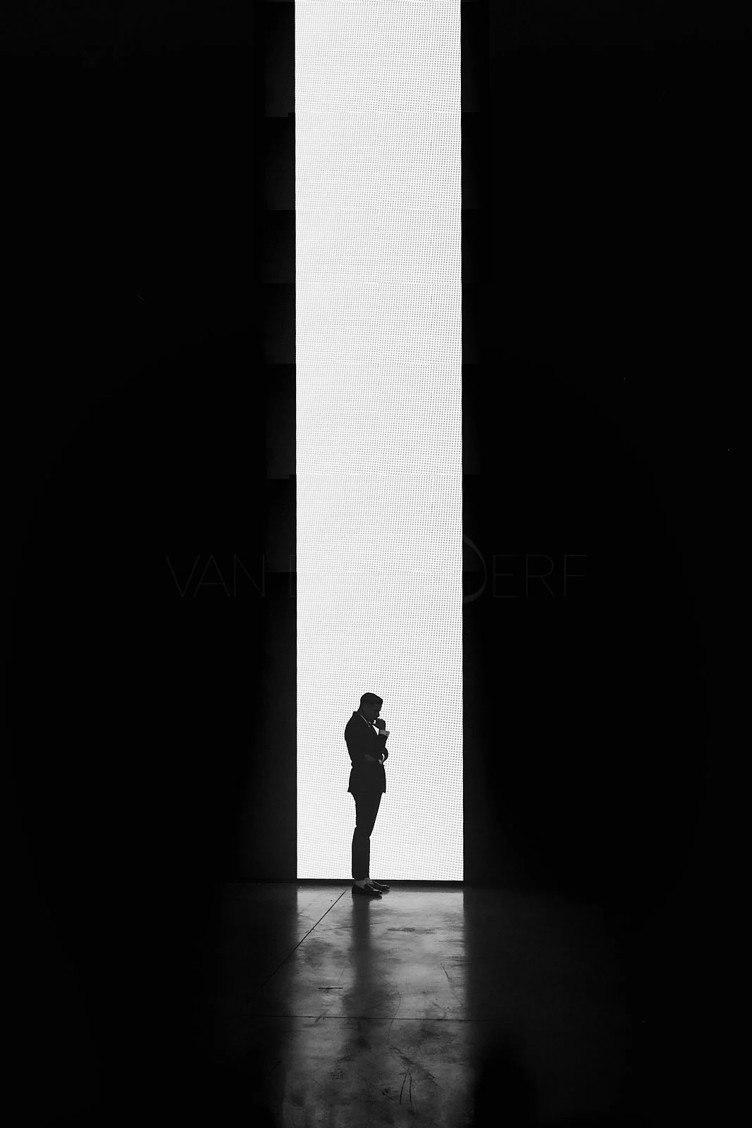 Silhouette of a man standing in front of a large vertical illuminated screen, with his hand on his chin, in a dark room.