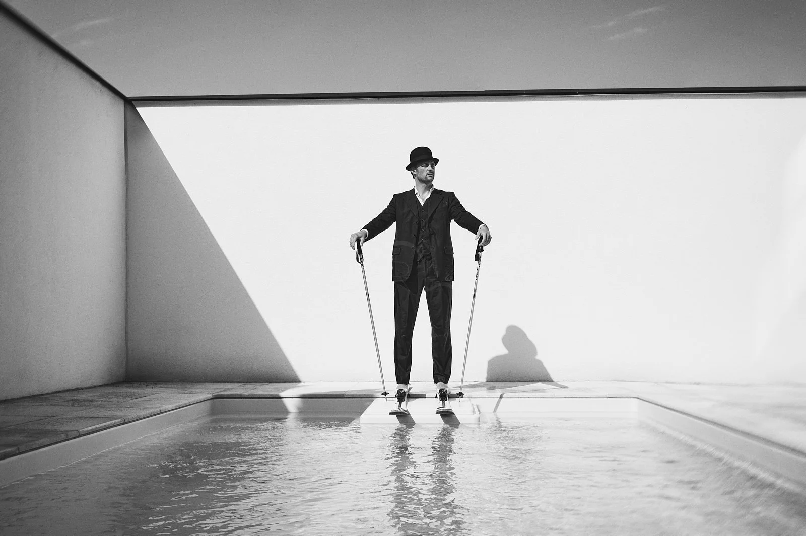 A man in a suit with a hat on stilts, standing on a small platform over water, with a minimalist white and gray background.