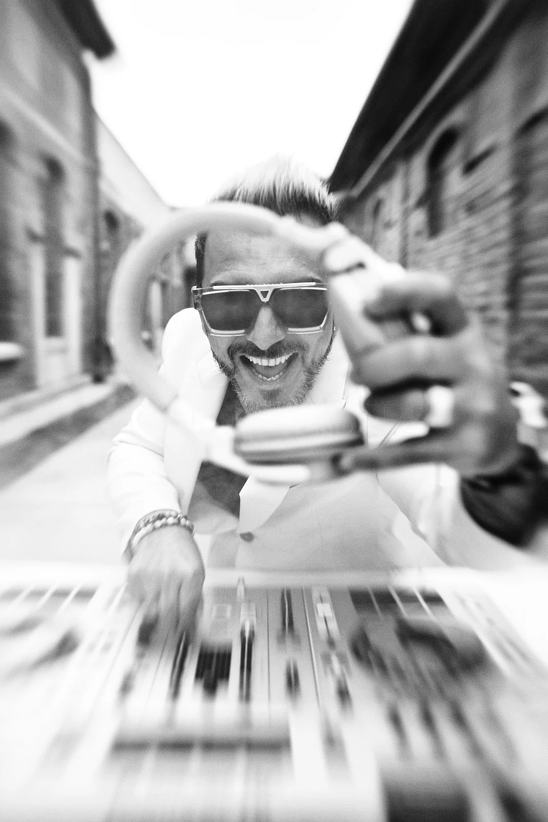 A smiling man with sunglasses leaning over a table, holding a pair of glasses close to the camera. The photo is taken from a low angle, with a blurred city street in the background.