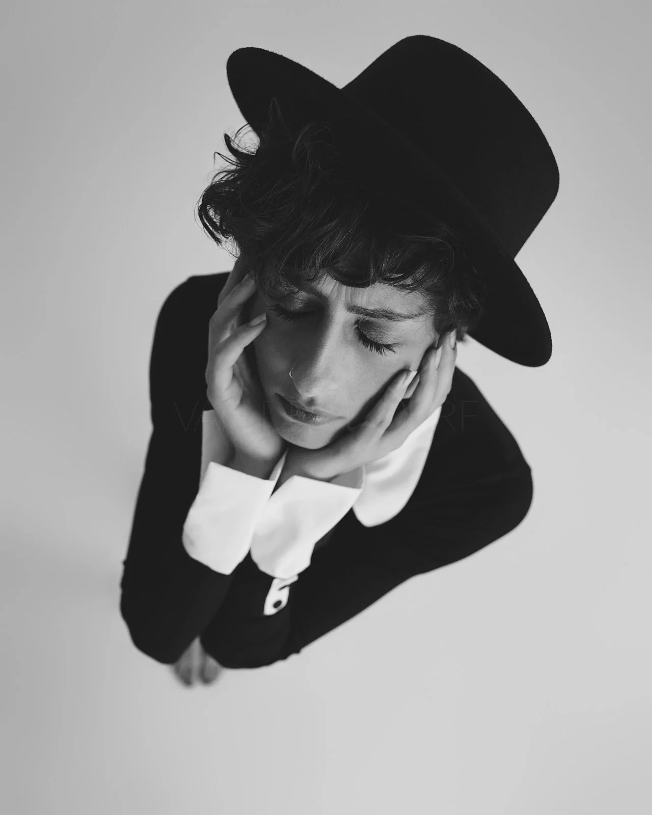 A black and white photograph of a woman wearing a black hat, with her hands touching her face and her eyes closed, expressing a sense of stress or contemplation.