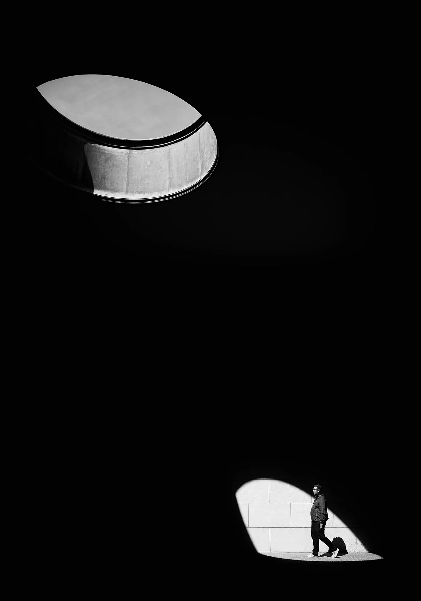 A person walking through a large dark space with a circular opening in the wall, casting light on the person and creating a shadow