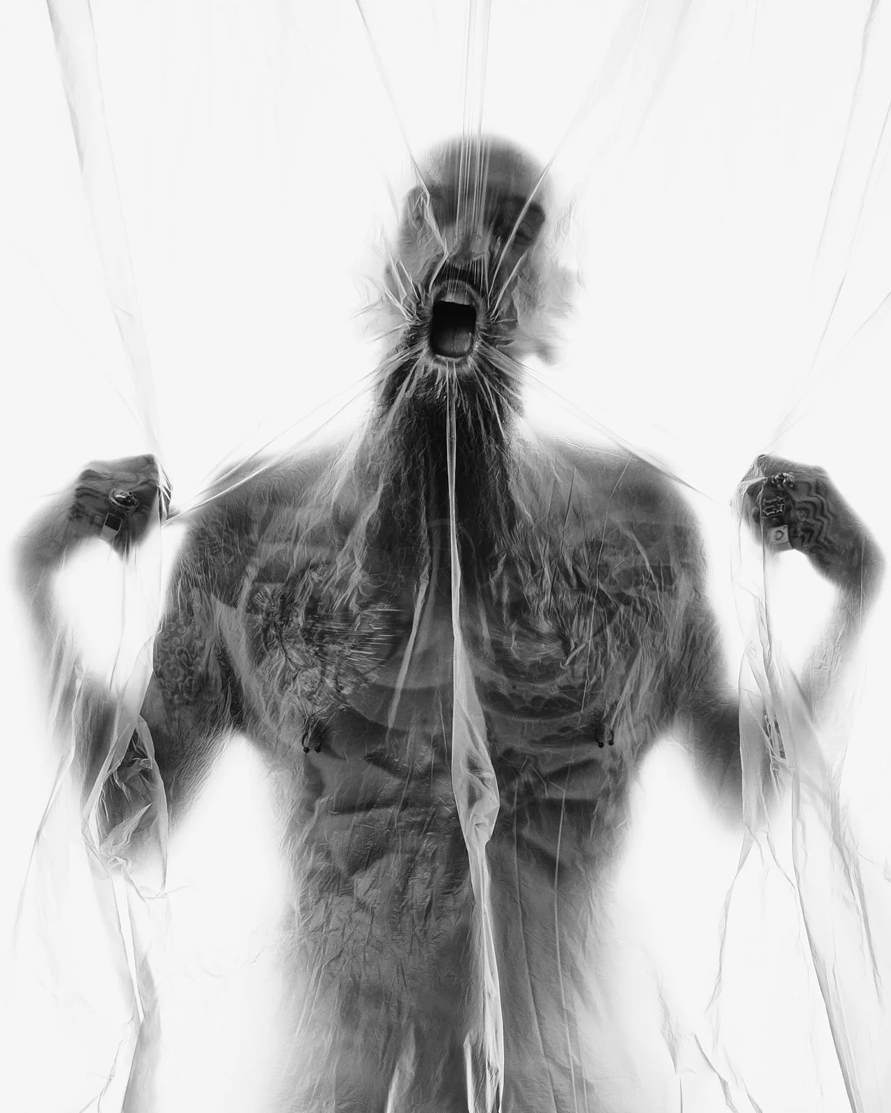 A man with tattoos and a beard posing behind crumpled transparent plastic, making a roaring face.