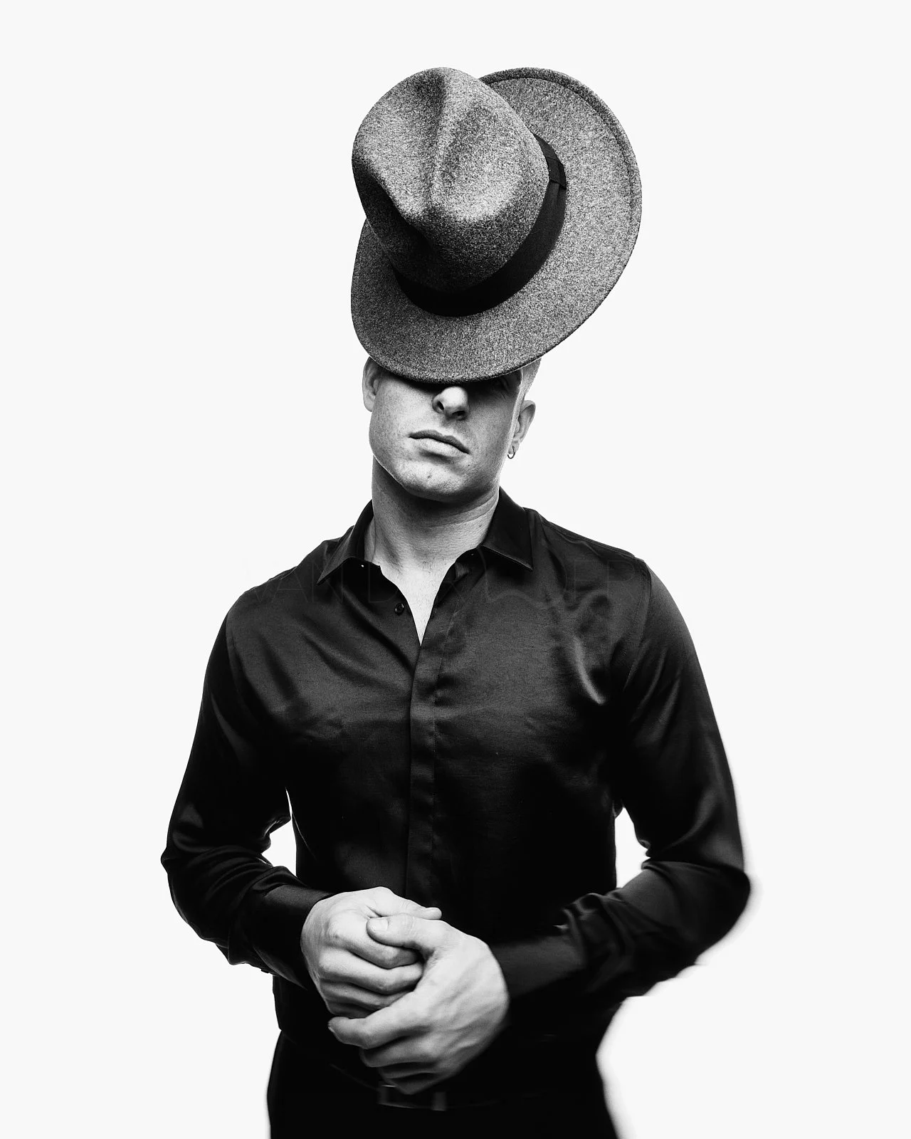 A black and white photo of a person wearing a black shirt with a large hat balanced on their forehead, obscuring their eyes, against a plain background.