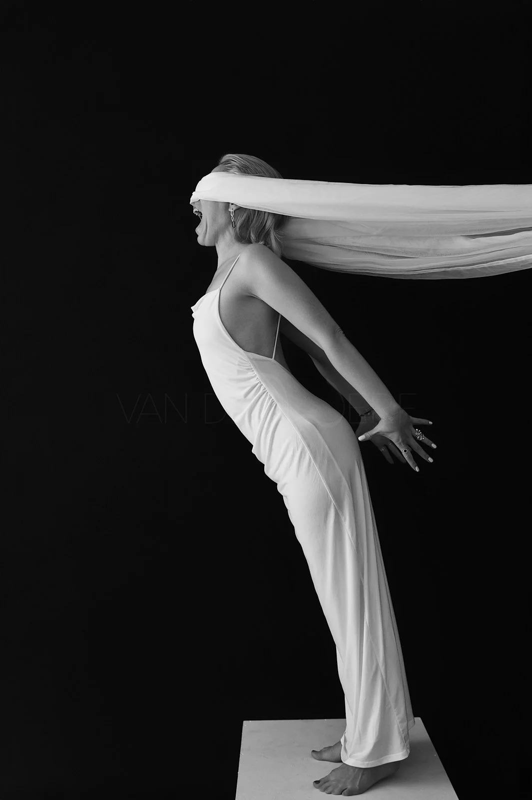 A woman in a white dress is posed on a platform against a black background, with her head turned to the side and a long white fabric flowing behind her.