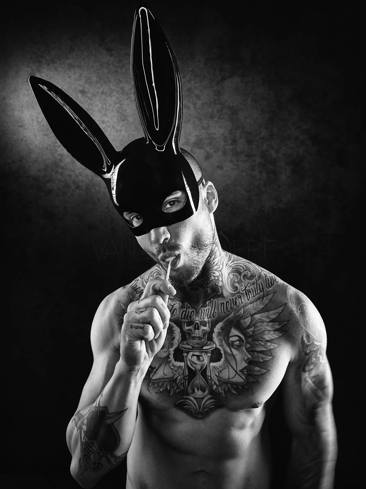 A shirtless man with tattoos on his chest and arms wearing a black bunny mask and licking a lollipop.