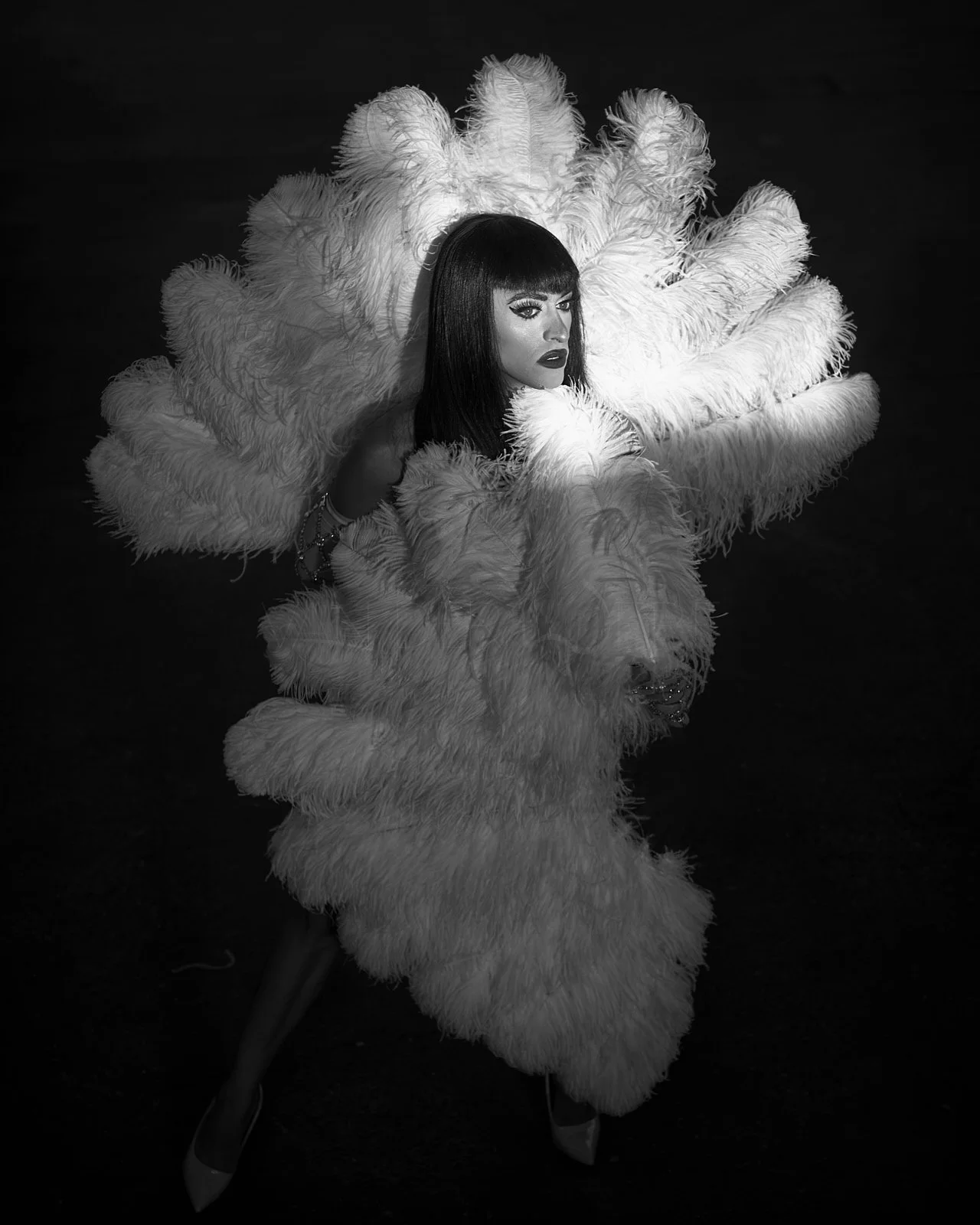 A woman with dark hair and bangs wearing dramatic makeup, posed inside large white feathered costume resembling a flower or bird, against a dark background.
