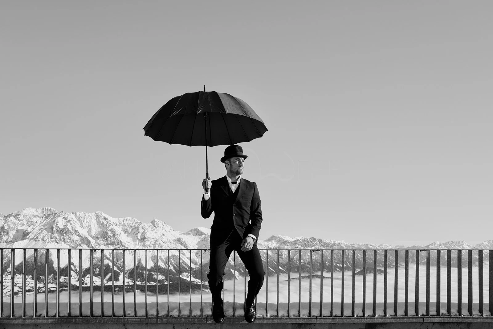 A man in a suit and hat holding an umbrella, standing on a ledge overlooking snow-capped mountains in the distance, in black and white.