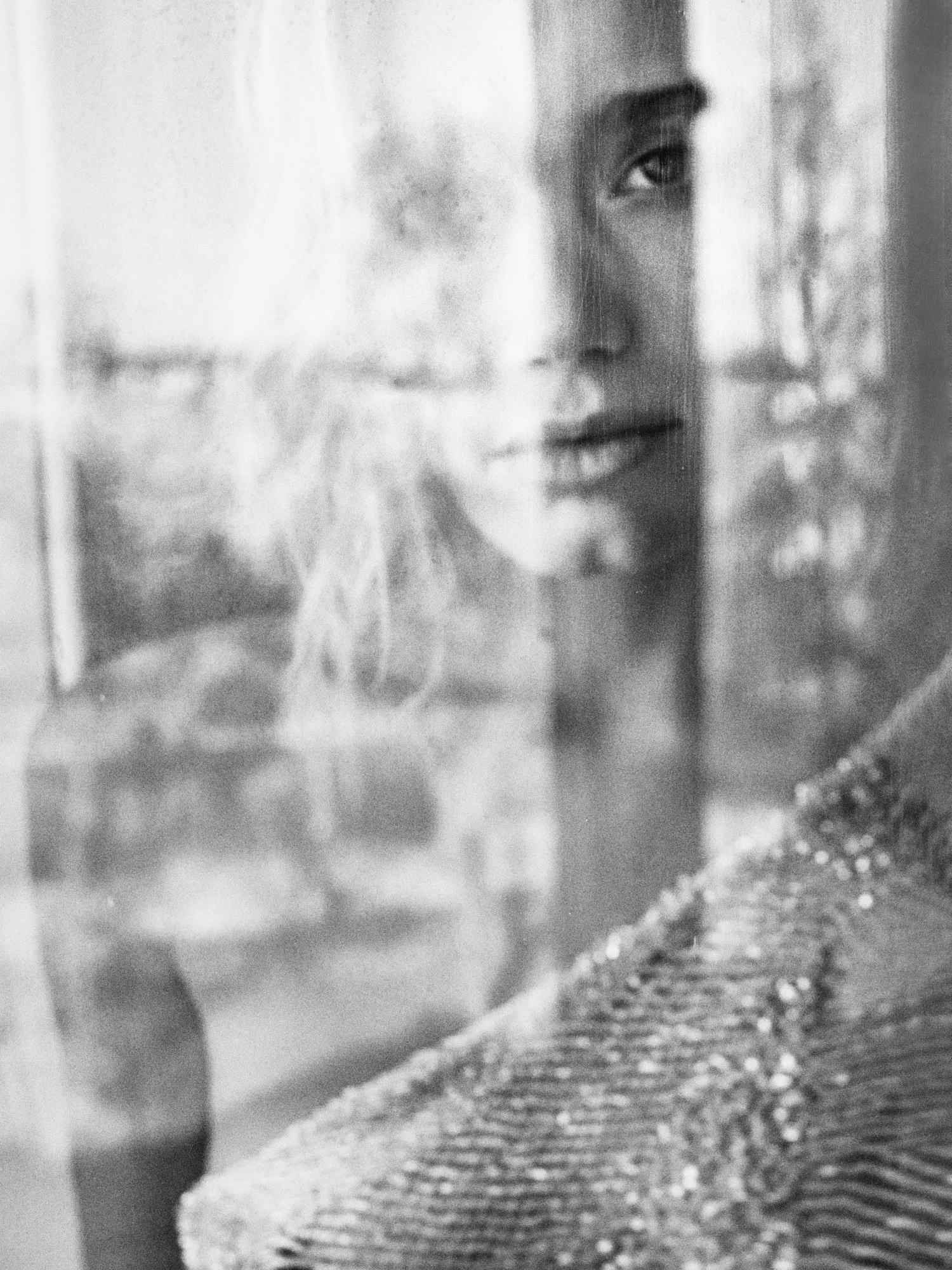 Black and white photograph of a woman seen through a glass surface, with her face slightly blurred and partly obscured by reflections and textures.