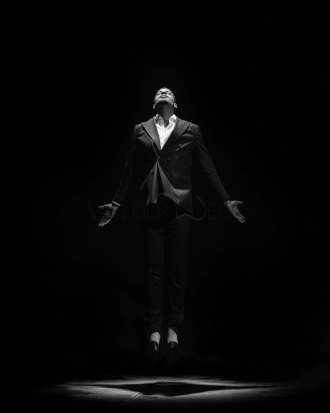 A man in a suit appears to be levitating with arms outstretched, facing upward, in a dark setting with a spotlight on him.