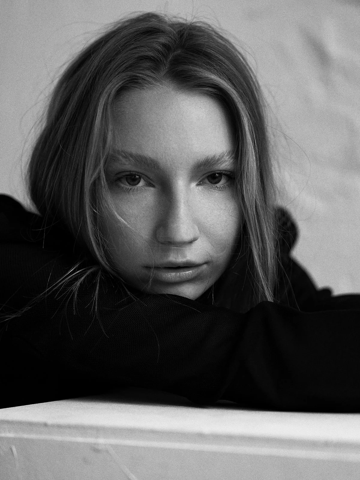 Close-up of a woman with light-colored hair, resting her head on her arms, looking directly at the camera in black and white.