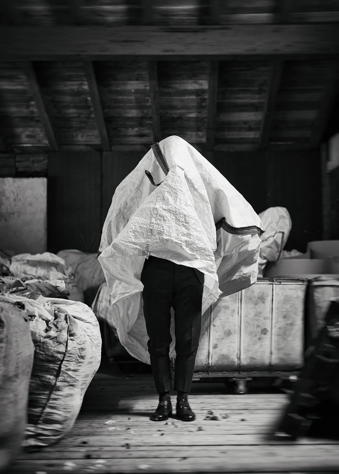 A person dressed in formal shoes and pants is carrying a large object wrapped in a white fabric or material, obscuring their face and upper body, in an industrial or warehouse setting with bags or sacks on the sides.