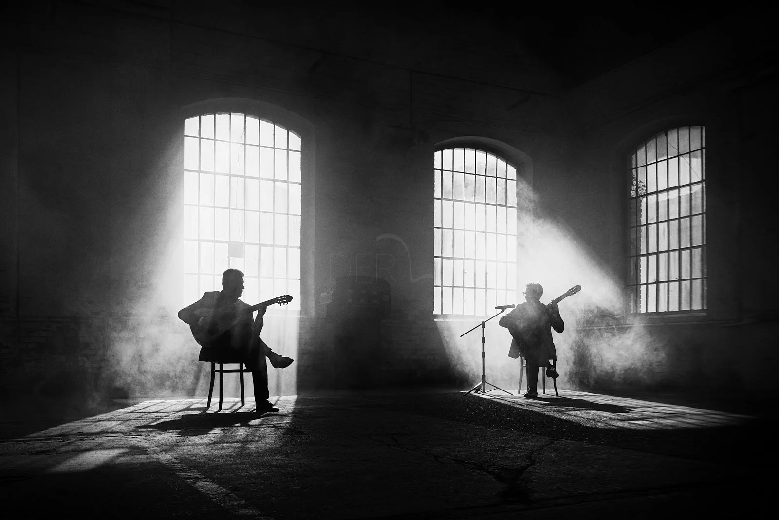 Black and white photo of two guitarists performing in a dimly lit space with large industrial-style windows and fog, creating a moody atmosphere.