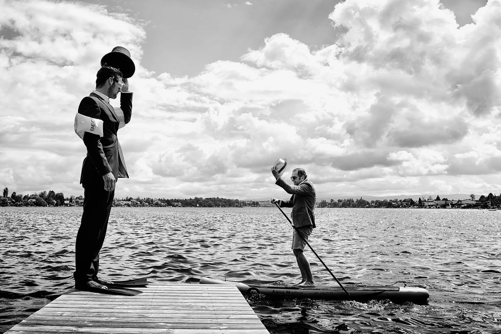 A man in a suit standing on a dock, holding a phone to his ear, while another man in casual clothes stands on a paddleboard near the dock, tipping his hat, on a lake with cloudy sky.