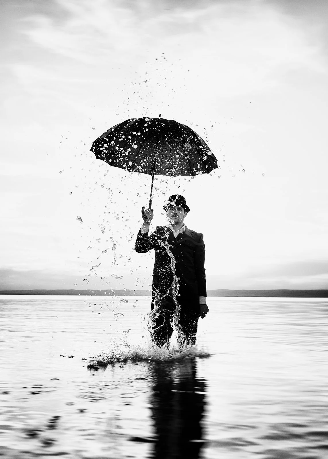 A person in a suit holding an umbrella standing in water, with splashes around, in a black and white photo.