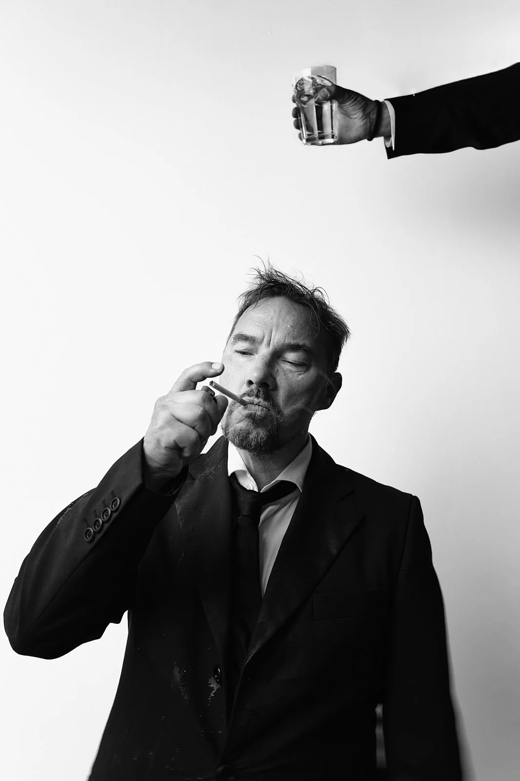 Black and white photo of a man in a suit smoking a cigarette, with a hand holding a glass of liquor extended from the top right corner.