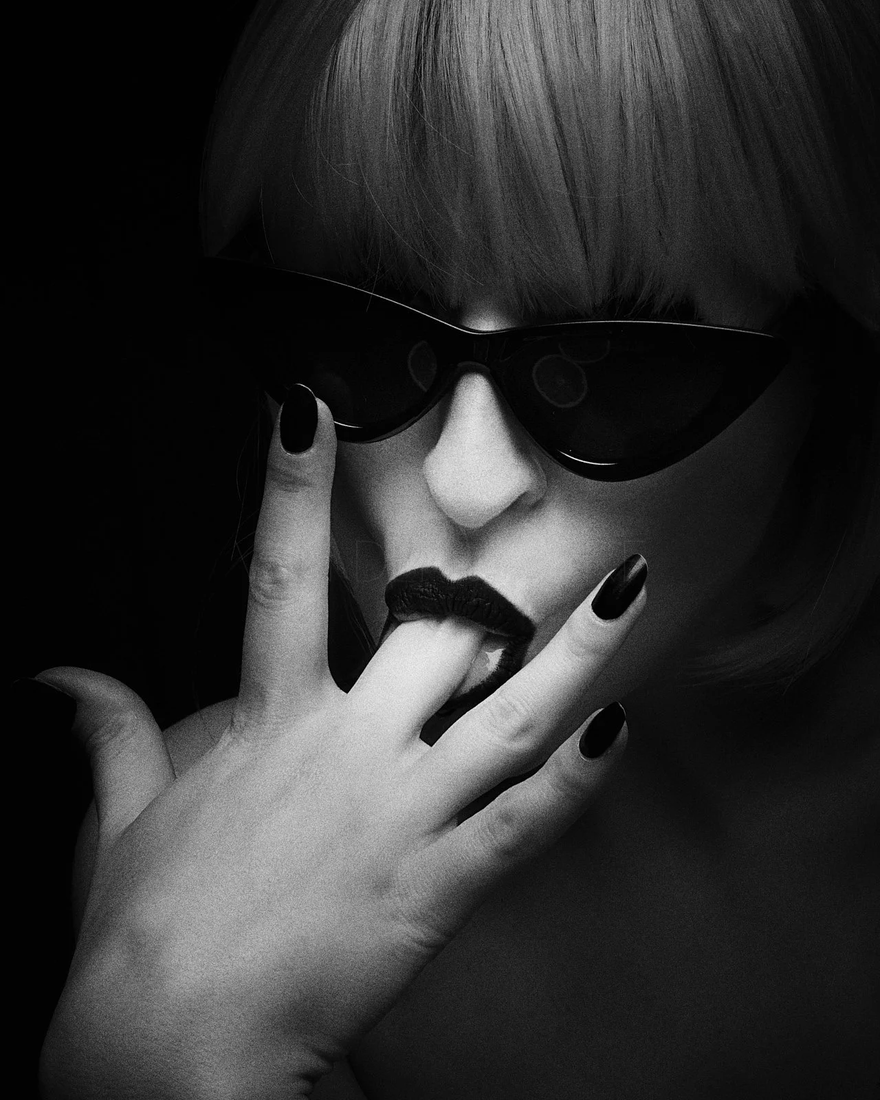 A black and white photo of a woman with short hair, wearing sunglasses and dark lipstick, touching her lips with her fingers.