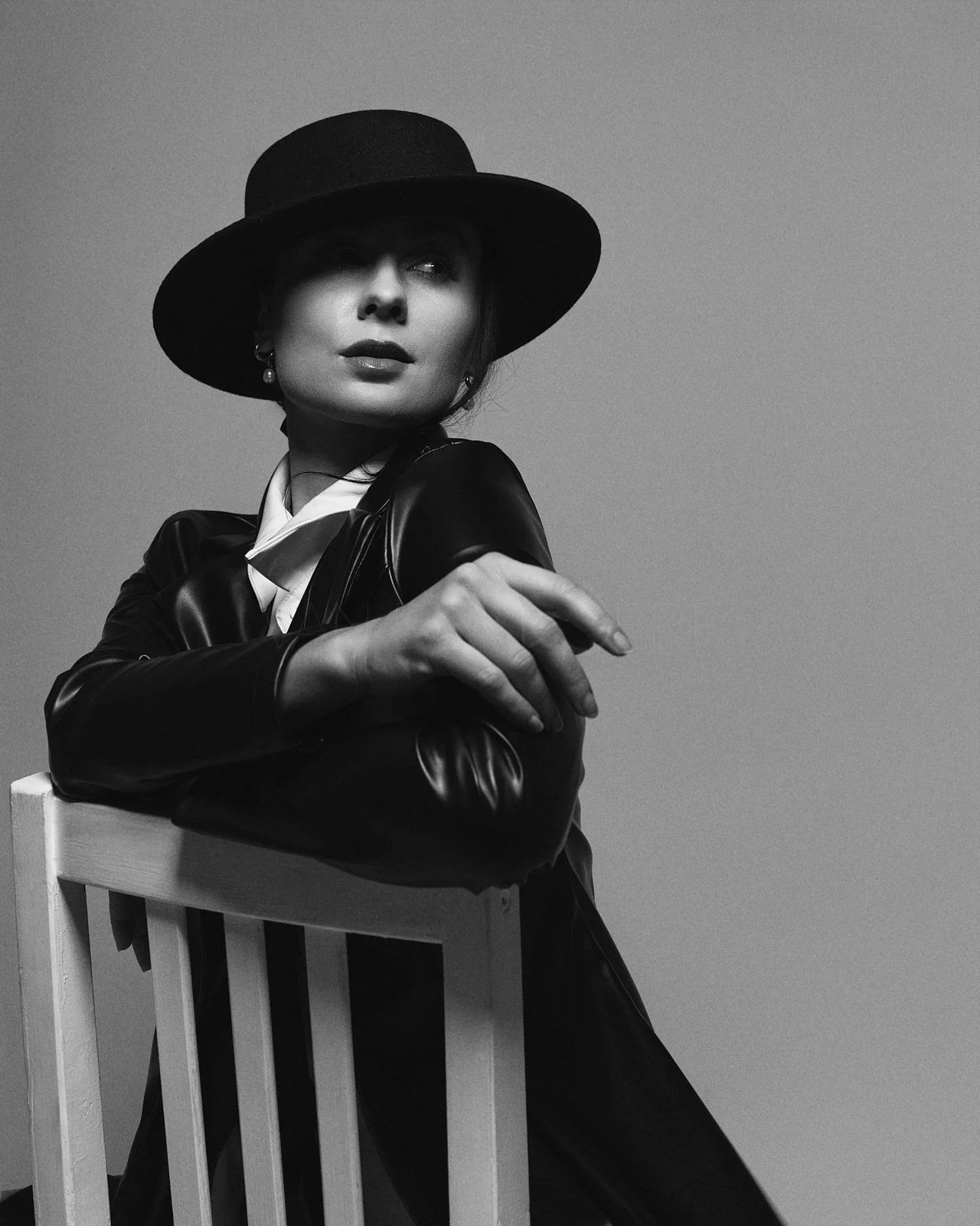 A woman wearing a wide-brimmed hat and leather jacket sitting on a chair in black and white photo.