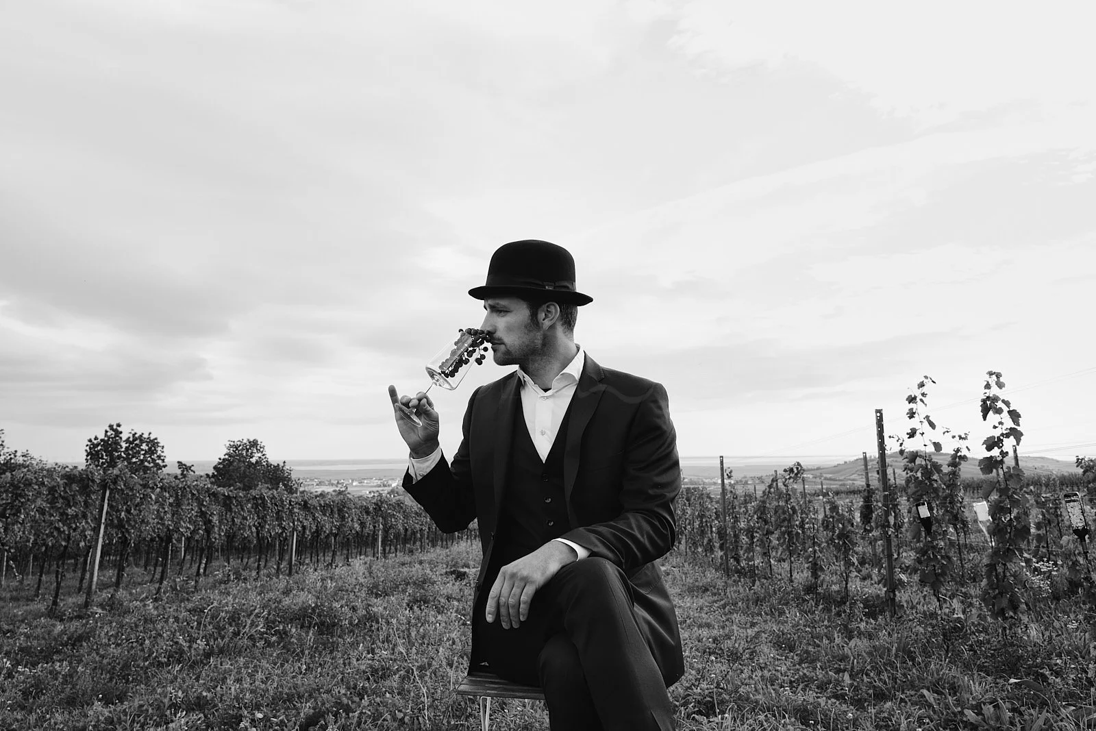 Man in a suit and hat sitting in a vineyard, drinking wine from a glass, in black and white.